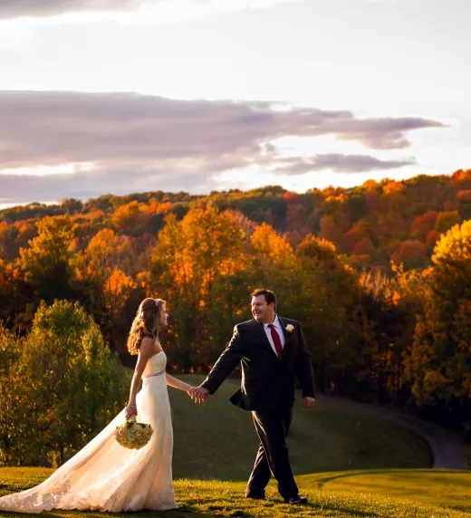 Bride and groom walking hand in hand with fall foliage behind them.