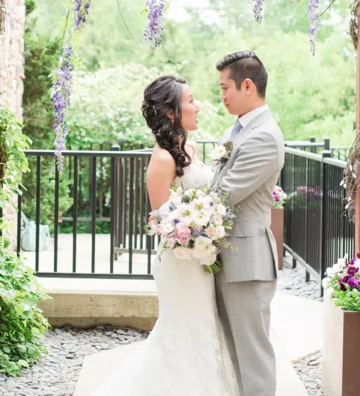 Bride and groom stand underneath purple hanging flowers.
