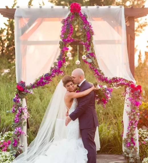 Groom kisses bride under their floral ceremony arch.
