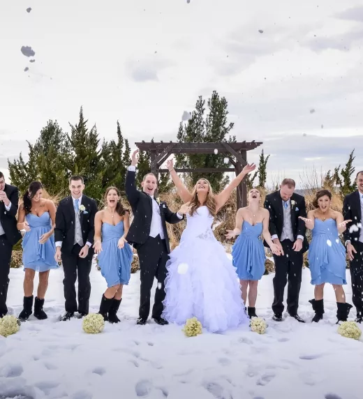 Wedding party, featuring men in black tuxes and bridesmaids in short blue dresses, stand together in a line and throw snow in air.