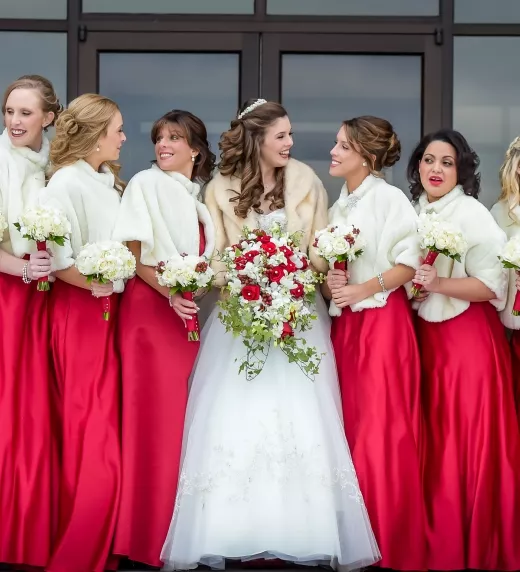 Bridesmaids, wearing long red dresses and white furry shawls, stand with bride.