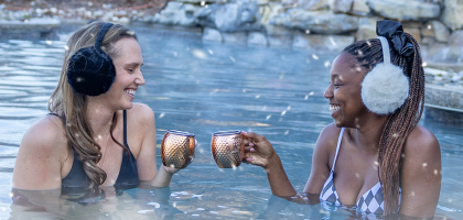 Two women in outdoor snowpool wearing earmuffs and drinking mules.