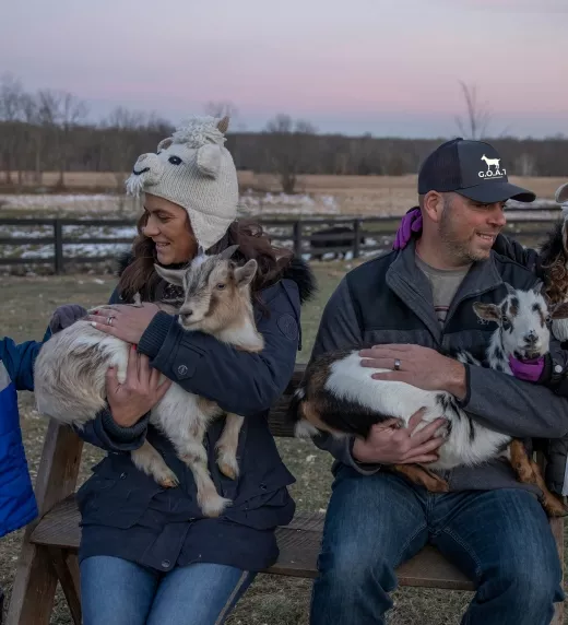 Family of four wearing goat hats and holding baby goats.